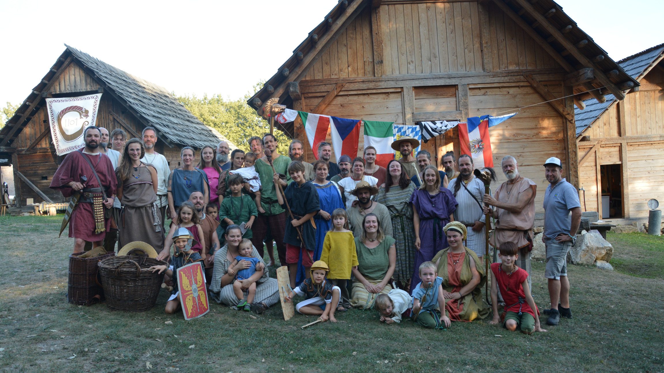 Gruppenfoto einer Gruppe von Menschen, die in mittelalterlichem Gewand gekleidet sind und auf einer Wiese vor Holzhütten steht,