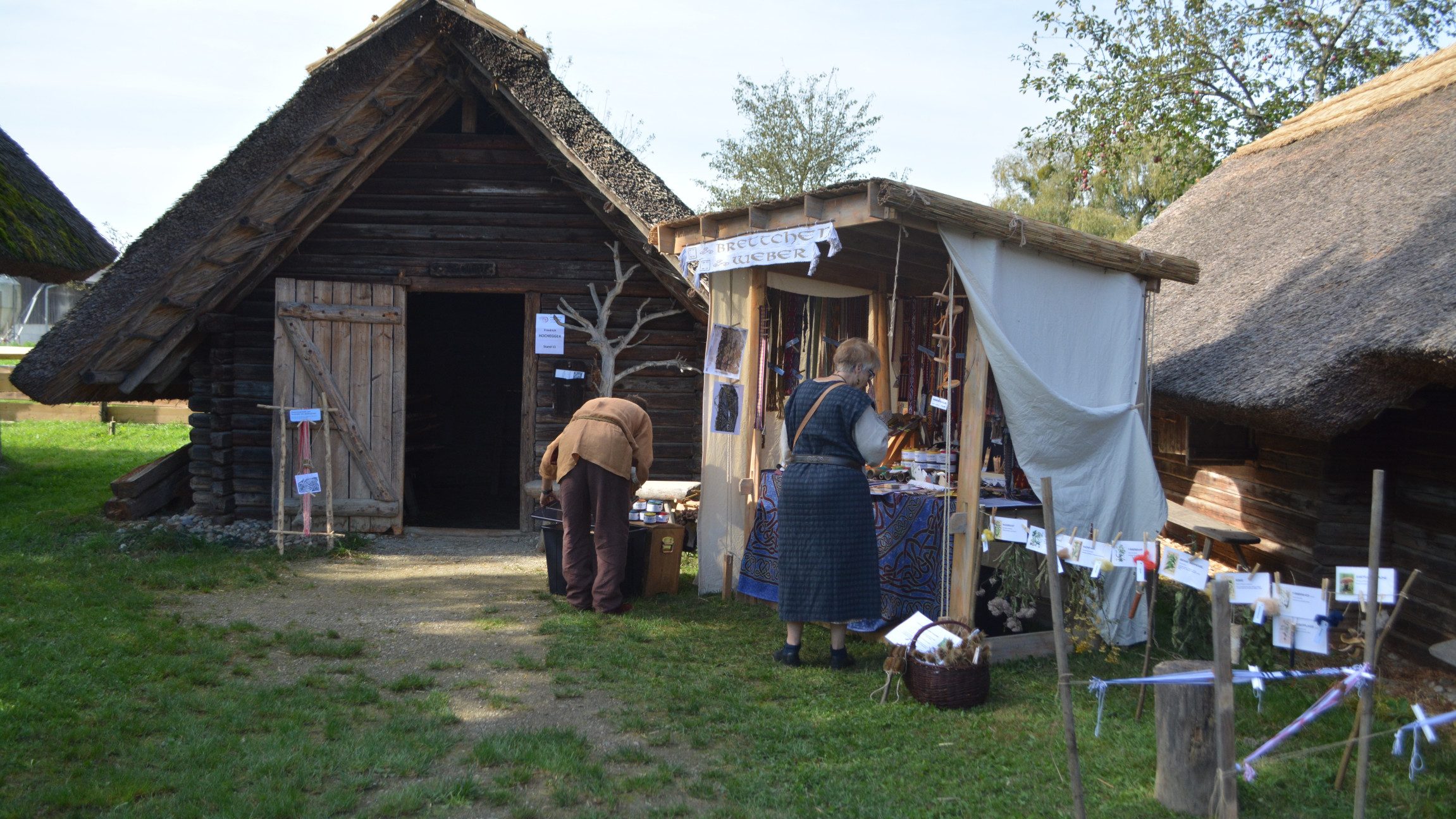 Blick auf einen Verkaufsstand, der neben einer Holzhütte steht, vor dem sich zwei mittelalterlich gekleidete Personen etwas ansehen.