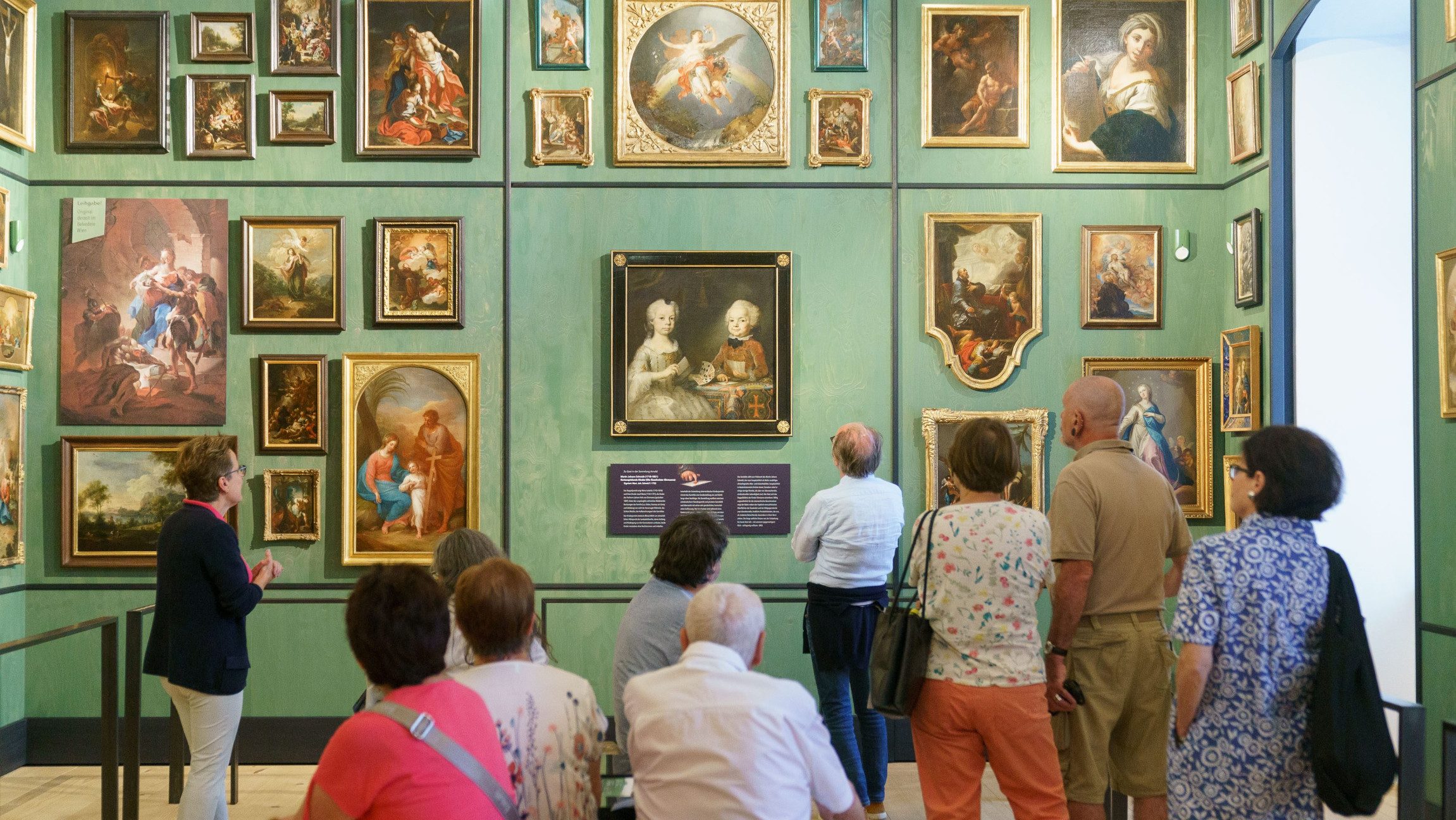 Foto von Menschen, die in einem Ausstellungsraum mit Gemälden stehen und sitzen und diese betrachten.