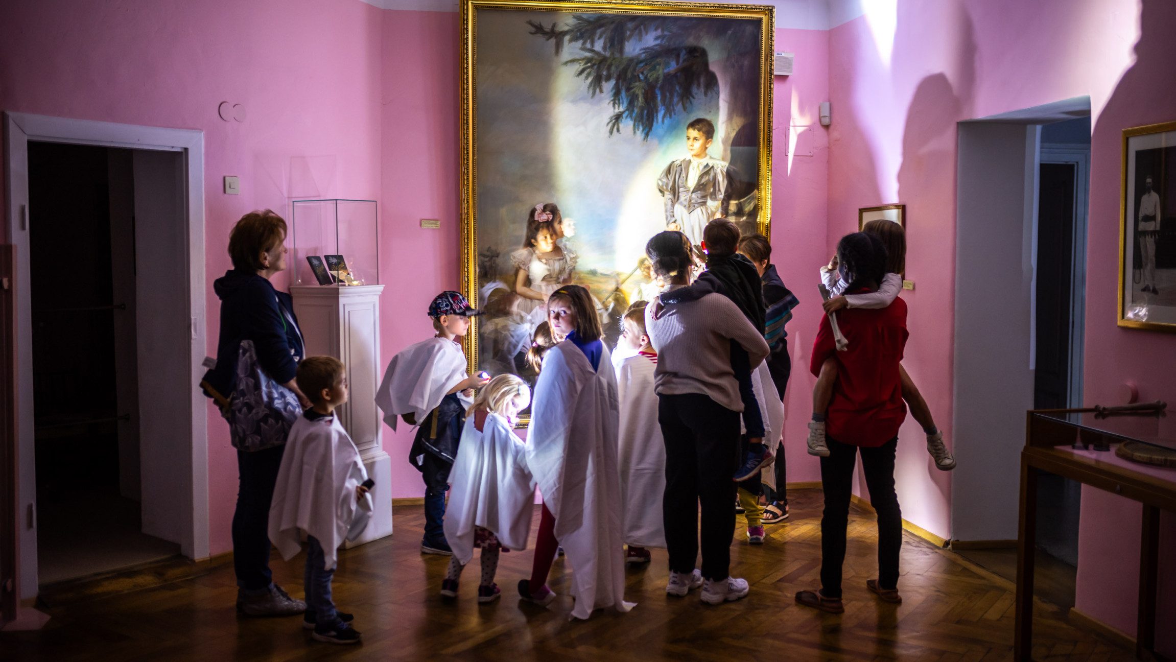 Foto von einer Gruppe Kindern, die in einem schwach beleuchteten Raum bei einer Führung vor einem Gemälde stehen.